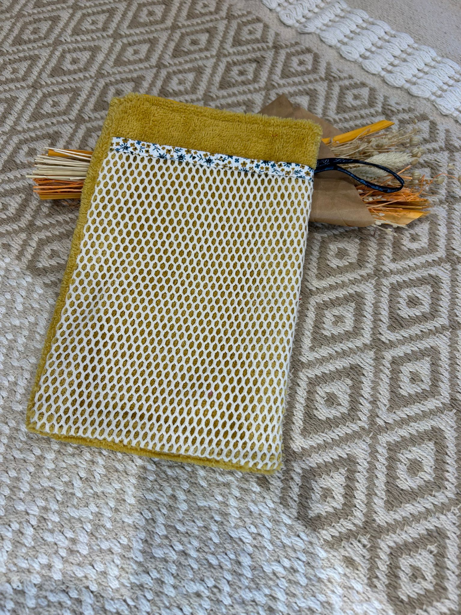 gant de toilette jaune avec filet blanc, sur un tapis beige avec des fleurs sécher 