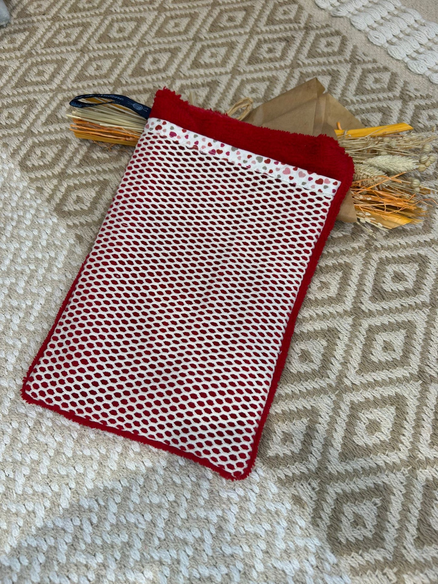 gant de toilette rouge, filet blanc et coeur sur un tapis beige et un bouquet de fleurs sécher 